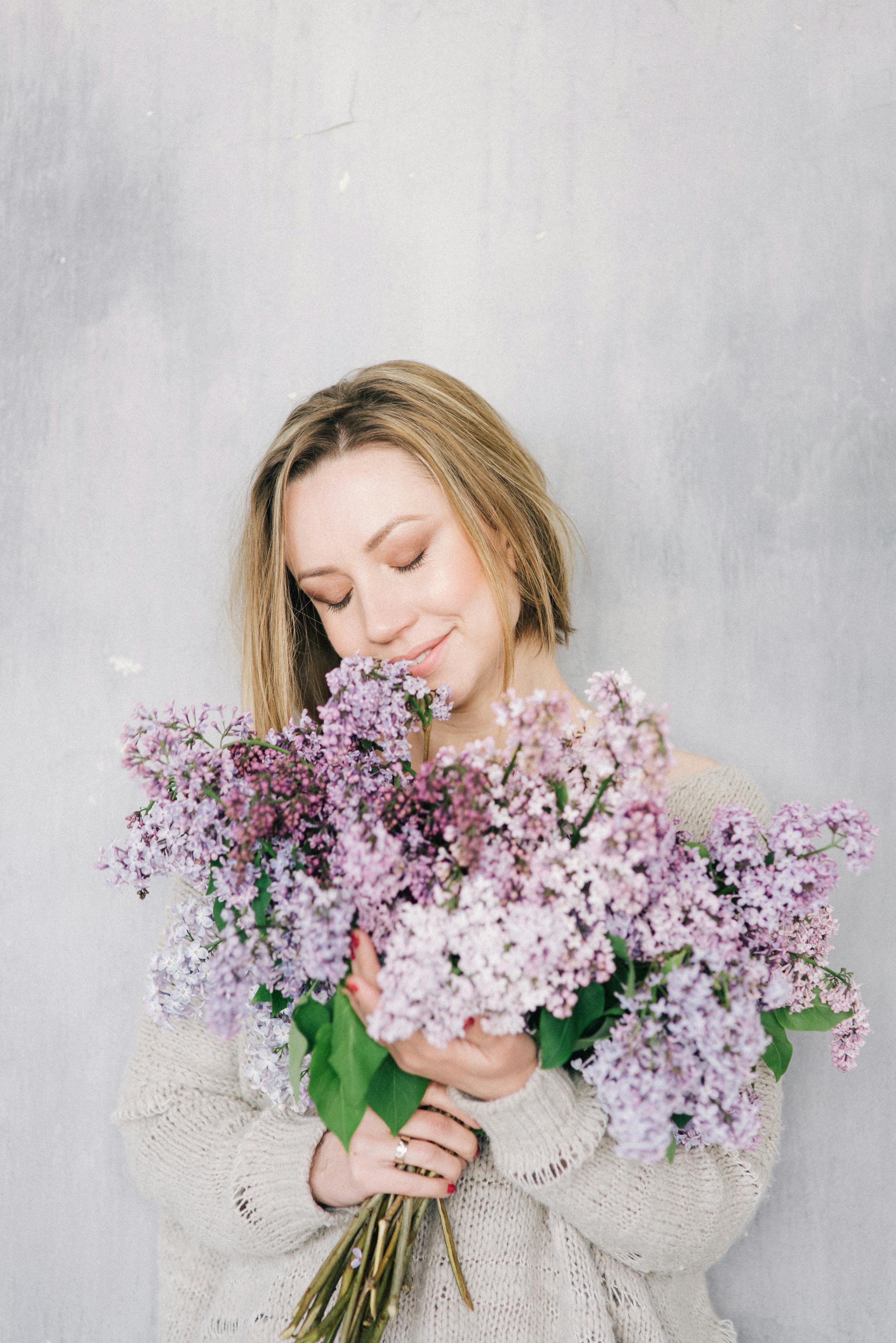 A Blonde Lady in Knitted Long Sleeves Holding Lilac Flowers